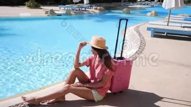 带粉红色箱子的年轻女子坐在酒店游泳池附近。 旅游度假概念。 高清视频支持1920x1080
